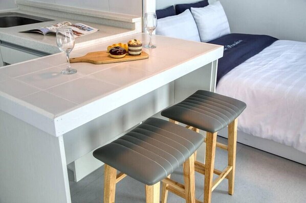 A compact kitchen area with a white countertop, two stools, a wooden cutting board with pastries, and a wine glass. - Jebudo Olle Pension (Hwaseong)
