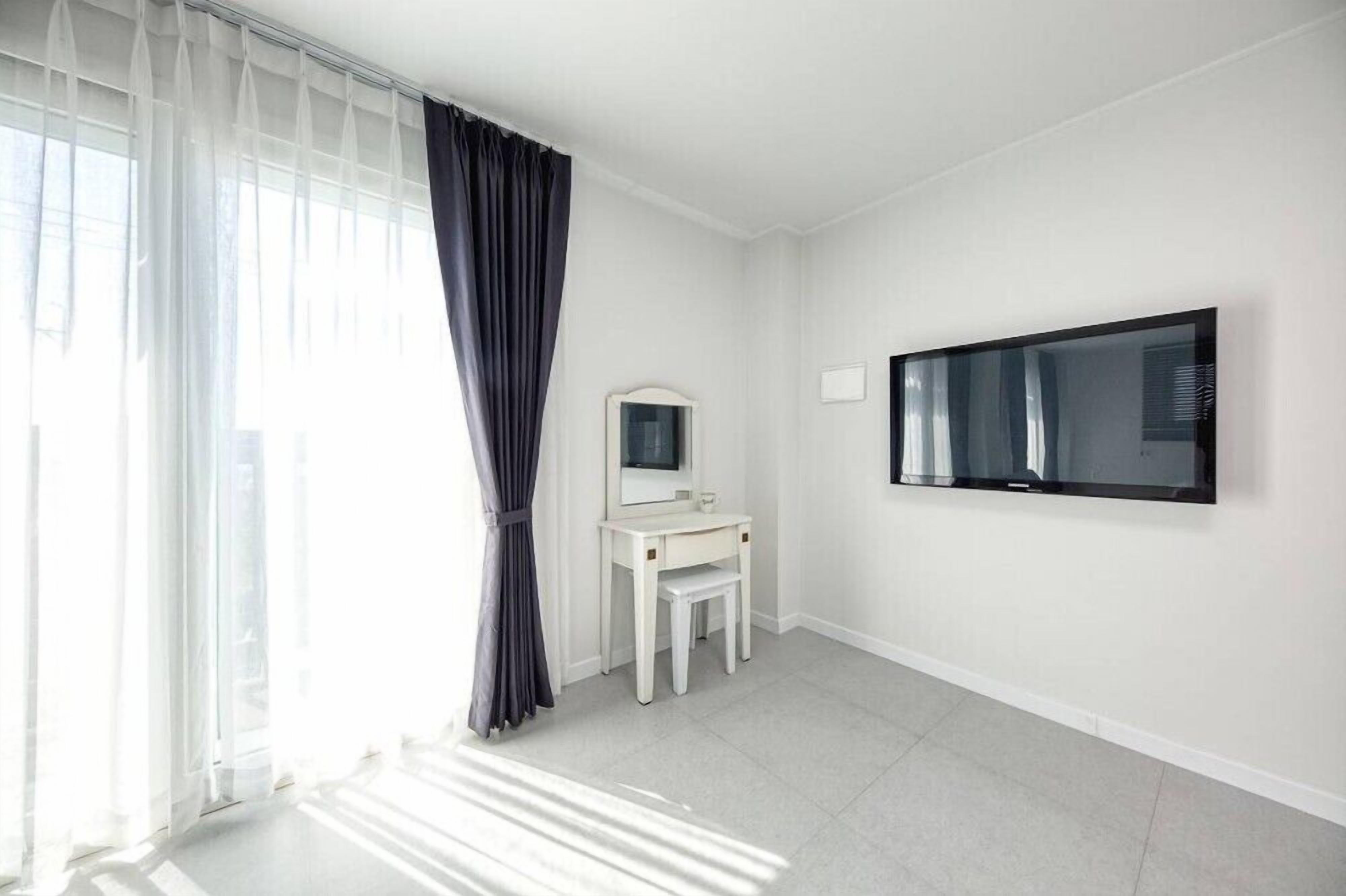 Ein heller, schlicht eingerichteter Raum mit einem großen Fenster, einem weißen Schminktisch mit Spiegel und einem an der Wand befestigten Flachbildfernseher.