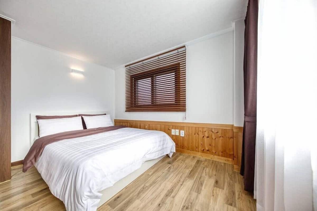 A bedroom with a wooden floor, a bed with white bedding, a wooden headboard, a window with blinds, and a wall-mounted light.