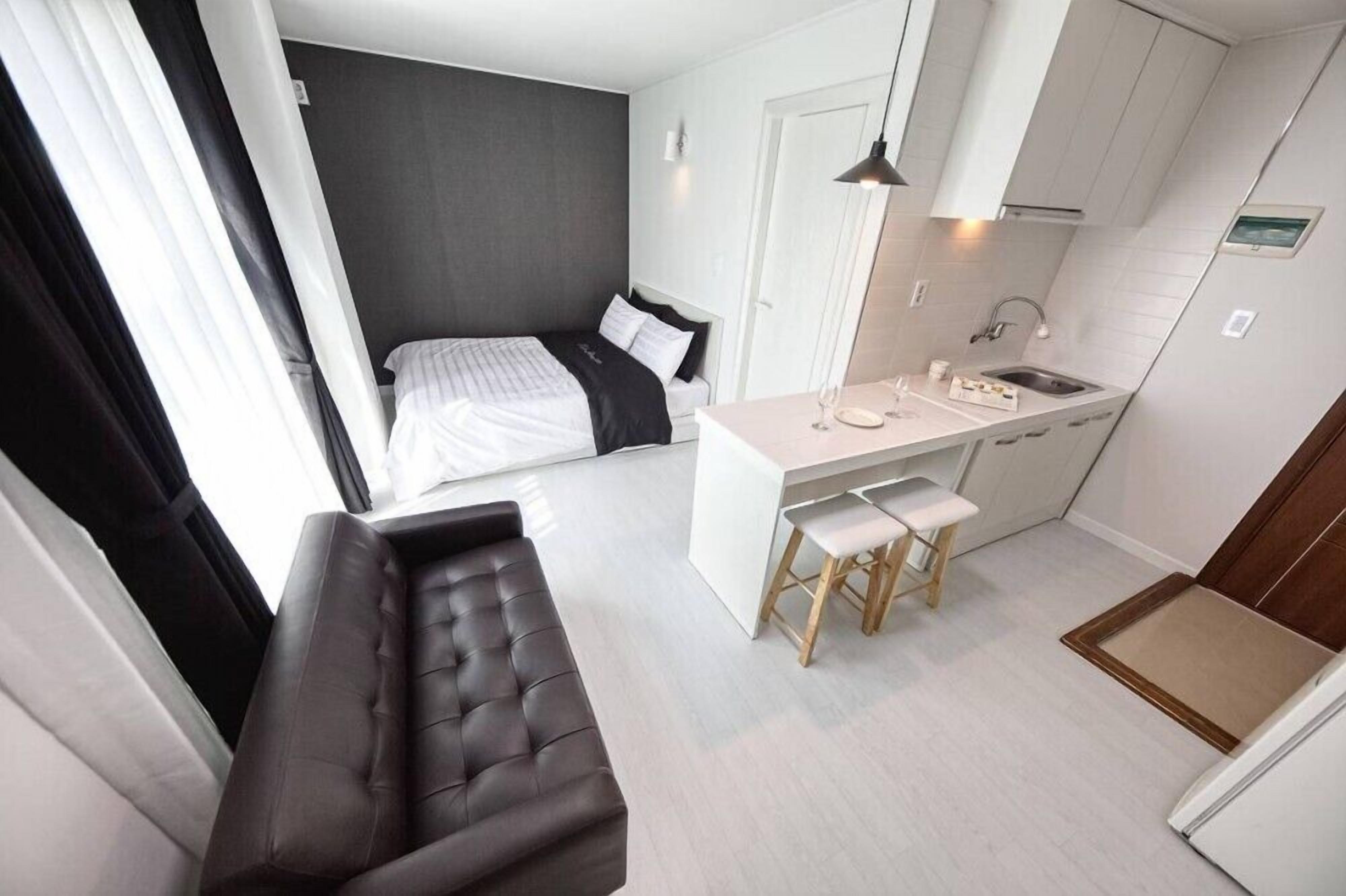 A compact room with a bed, a small kitchenette, a bench, and a window with curtains.
