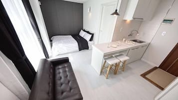 A compact room with a bed, a small kitchenette, a bench, and a window with curtains.