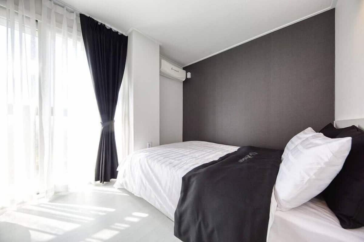 A modern hotel room with a large bed, white bedding, a dark headboard, and a window with sheer curtains.