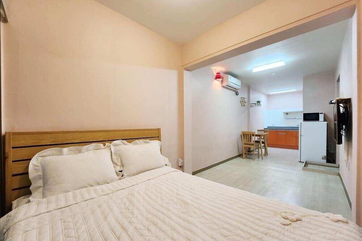Habitación de hotel con cama de cabecera de madera, una pequeña kitchenette con microondas y refrigerador, y un área de comedor con una silla y mesa.