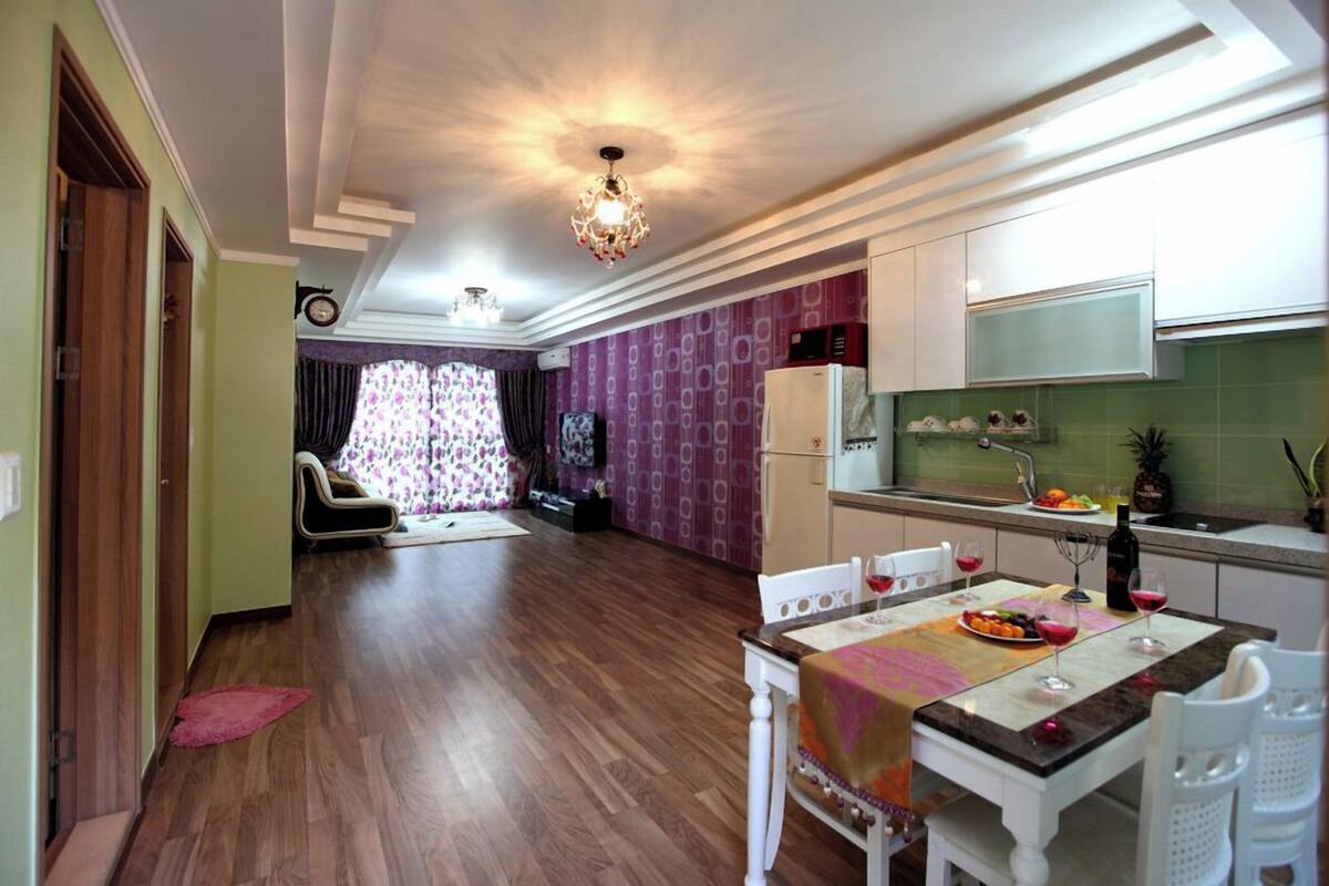 A modern kitchen with white cabinets, a dining area with a table set for two, and a living room with a sofa and curtains.