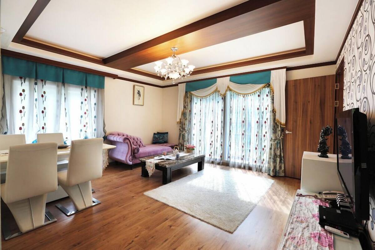 A spacious living room with a wooden floor, a dining area with chairs, a sofa, a coffee table, and curtains with a floral pattern.