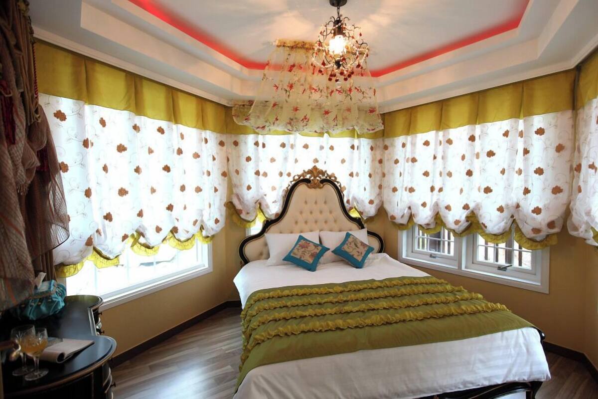 A bedroom with a large bed, floral curtains, a chandelier, and a view of the outdoors.