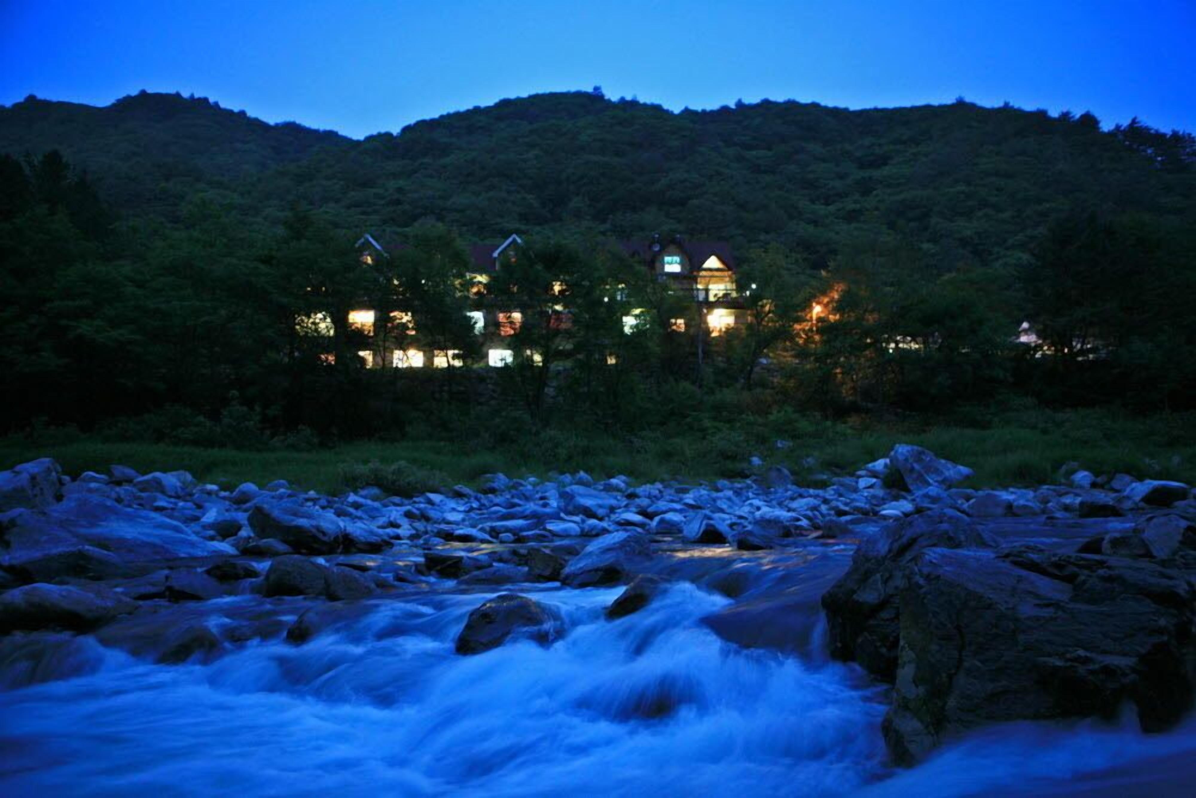 A riverside view of a hotel with multiple buildings, surrounded by lush greenery and a mountainous backdrop.