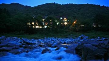 A riverside view of a hotel with multiple buildings, surrounded by lush greenery and a mountainous backdrop.