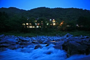 A riverside view of a hotel with multiple buildings, surrounded by lush greenery and a mountainous backdrop. - White Village Pension (Inje)