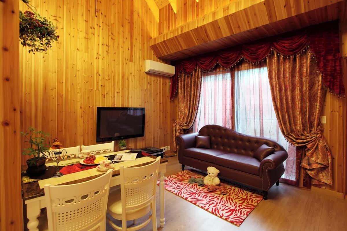 A cozy living room with a brown leather sofa, a flat-screen TV, wooden walls, and a dining area with a table and chairs.