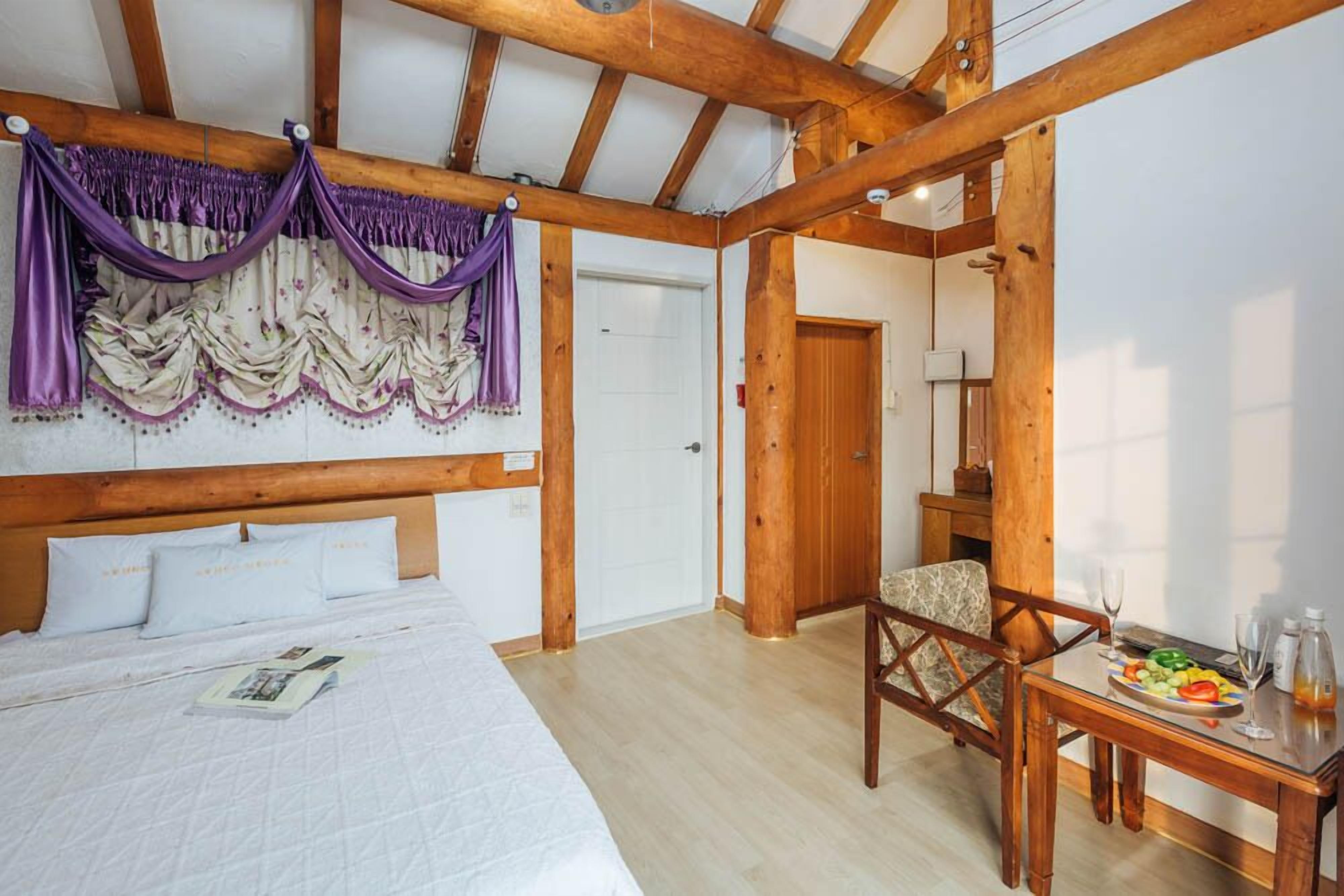 Un dormitorio con techo de madera, una cama con ropa de cama blanca, una silla, una mesa con una bandeja de frutas y una puerta.