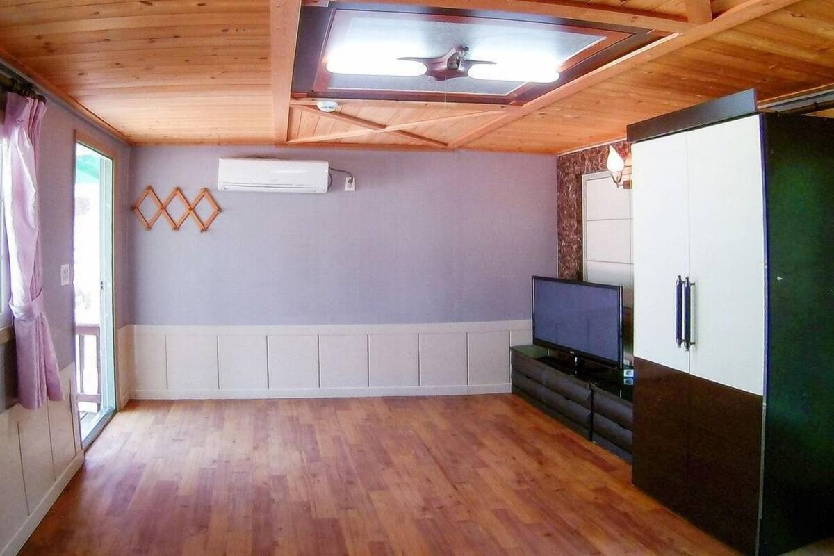 A room with wooden flooring, a ceiling fan, a window with curtains, a flat-screen TV, and a refrigerator.