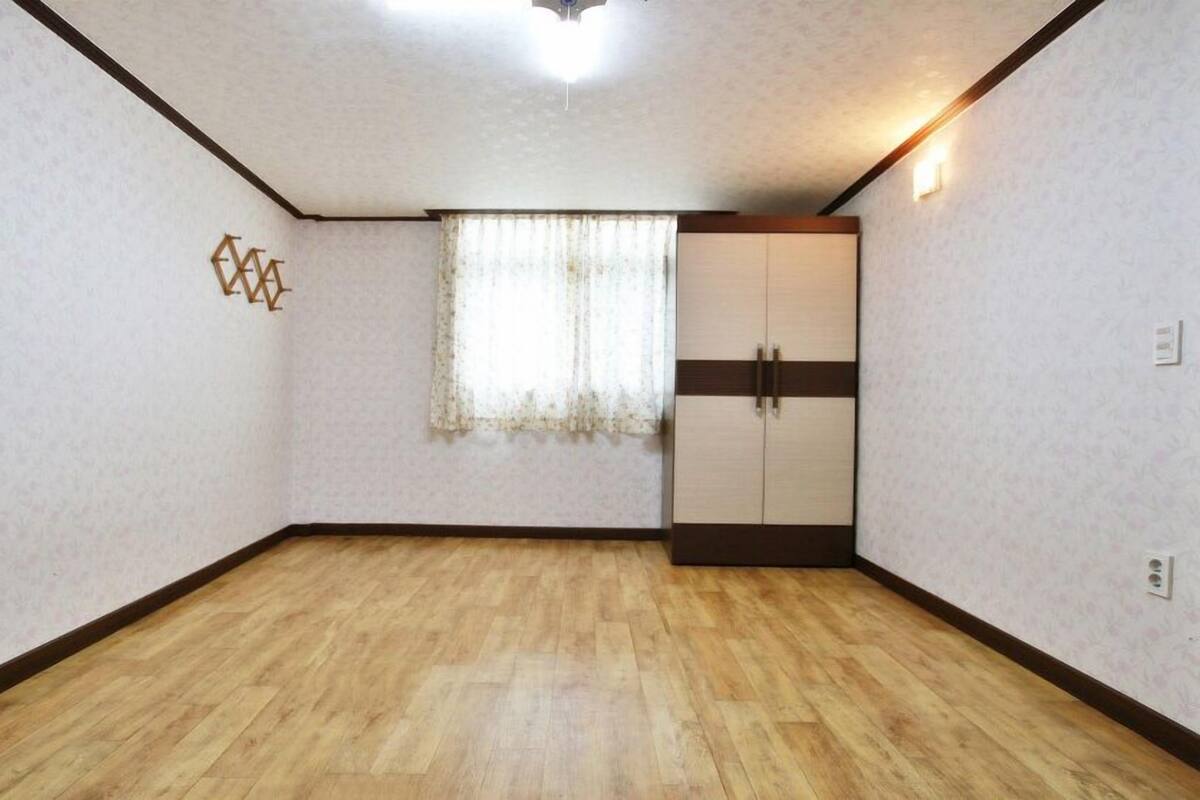 A room with wooden flooring, a window with lace curtains, a wardrobe with two doors, and a wall-mounted light fixture.