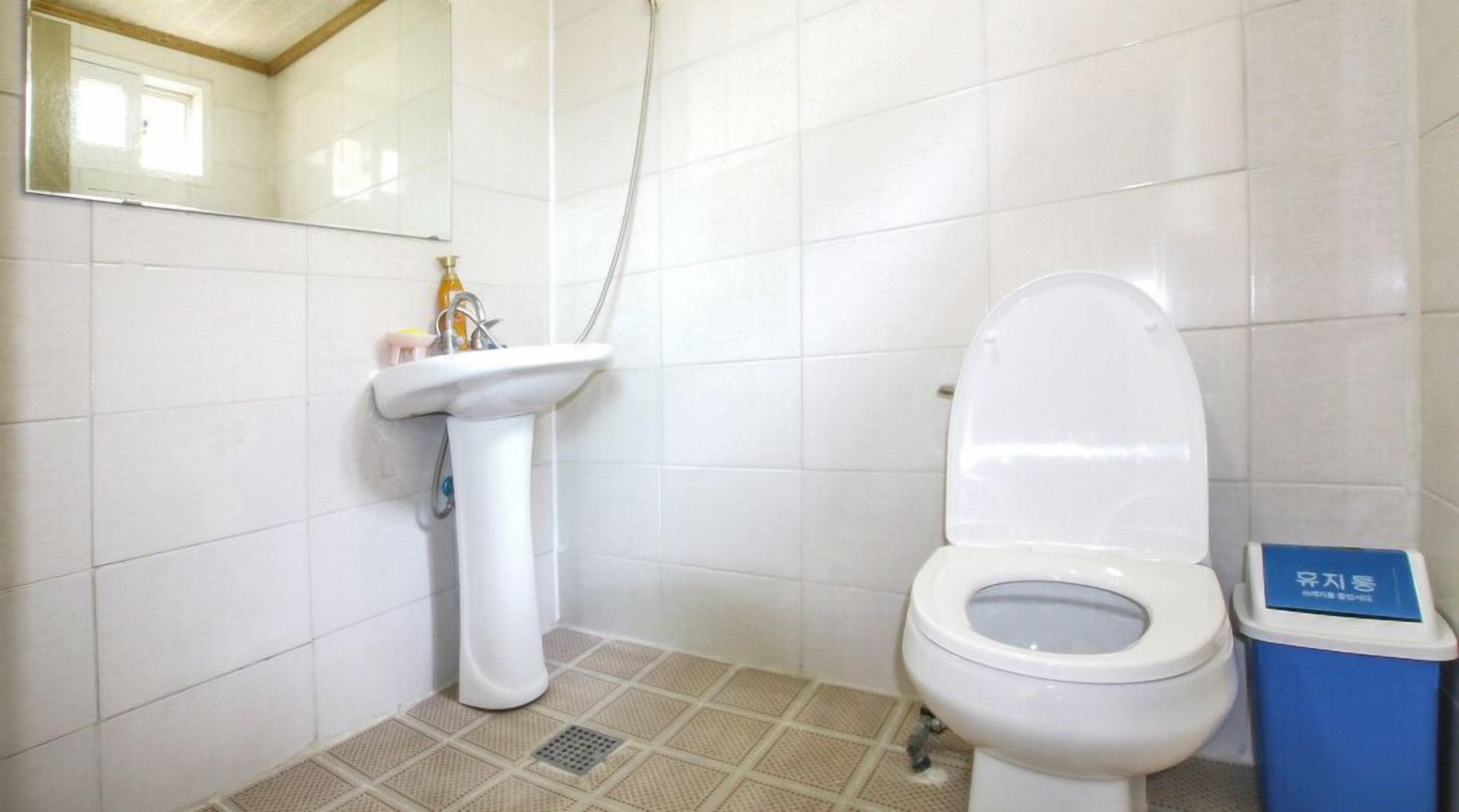 Une petite salle de bain propre, avec des carreaux blancs, un lavabo blanc, des toilettes et une poubelle bleue.