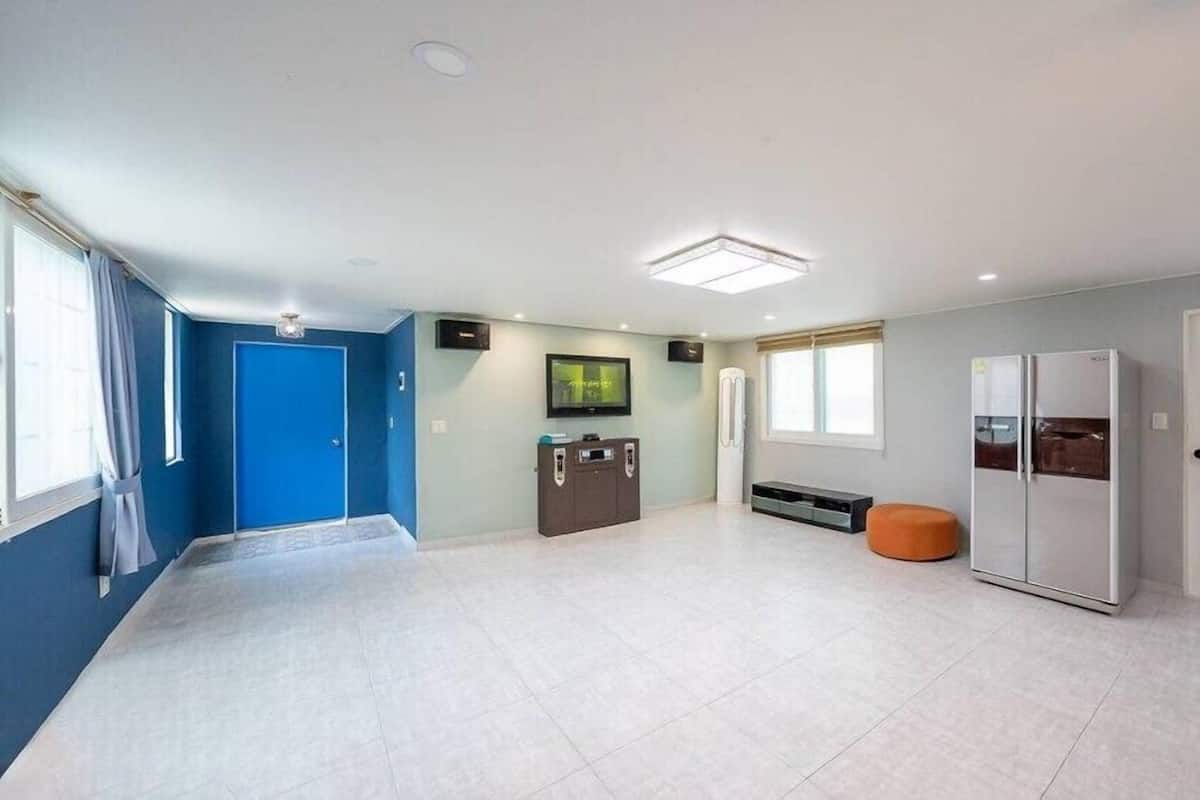 A spacious room with a blue door, a white refrigerator, a TV, and a bench.