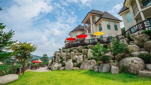 A multi-story building with a stone retaining wall, outdoor seating with umbrellas, and a clear sky. - Wellbeing log Pension (Gapyeong)