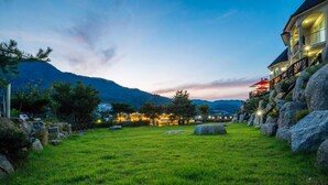 A well-maintained grassy area with large rocks, a building with multiple balconies and outdoor lighting, and mountains in the background. - Wellbeing log Pension (Gapyeong)