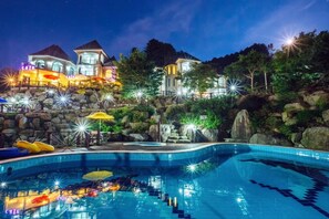 A swimming pool with a stone wall and a view of a large house with multiple levels and lights. - Haneul mok Pension (Gapyeong)