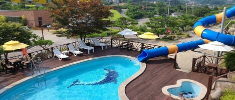 A swimming pool with a slide, surrounded by lounge chairs and umbrellas, set against a backdrop of greenery and hills.