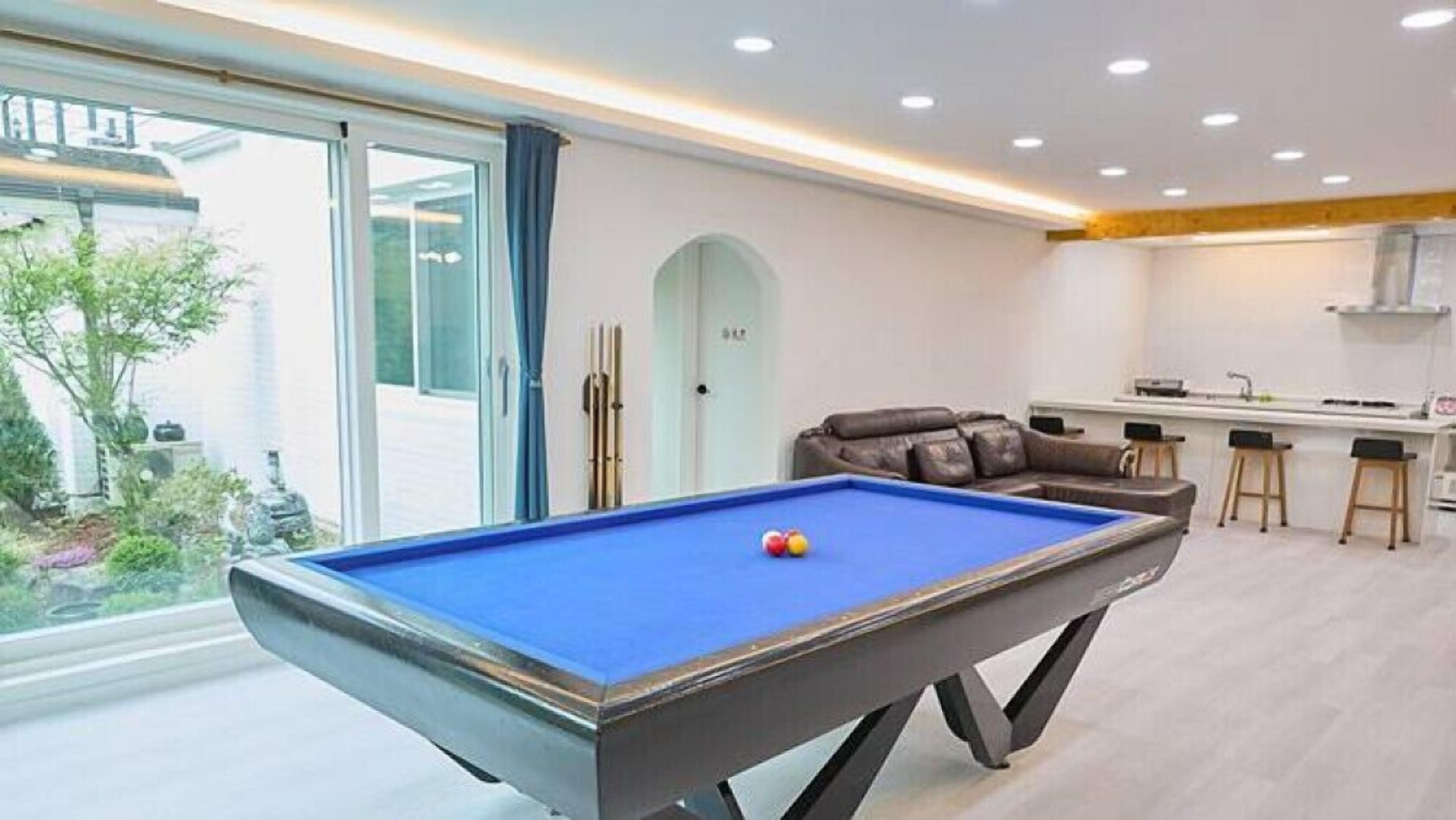 Una mesa de billar en una habitación con zona de cocina, un sofá y vistas a un jardín.