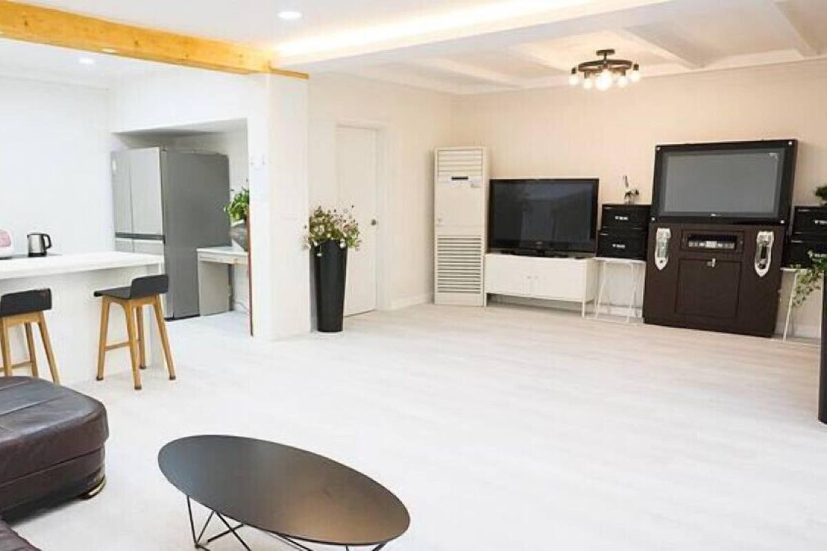 A modern living room with a sofa, a dining area with stools, a flat-screen TV, and a kitchen area with a refrigerator and a microwave.