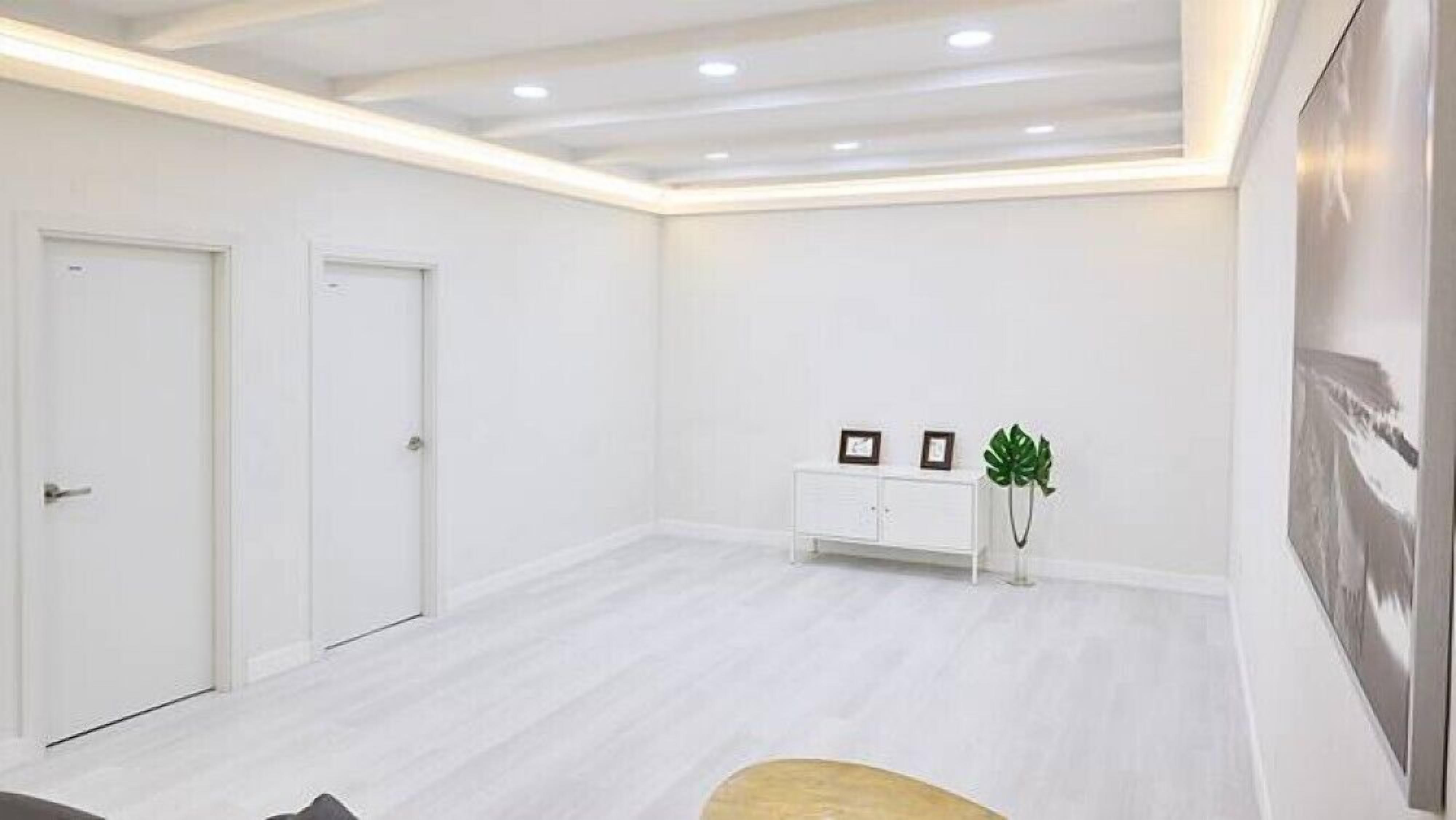 Una habitación espaciosa con paredes blancas y suelo de madera clara. Hay una consola blanca con dos cuadros enmarcados y una planta verde. Se ven dos puertas blancas a la izquierda.