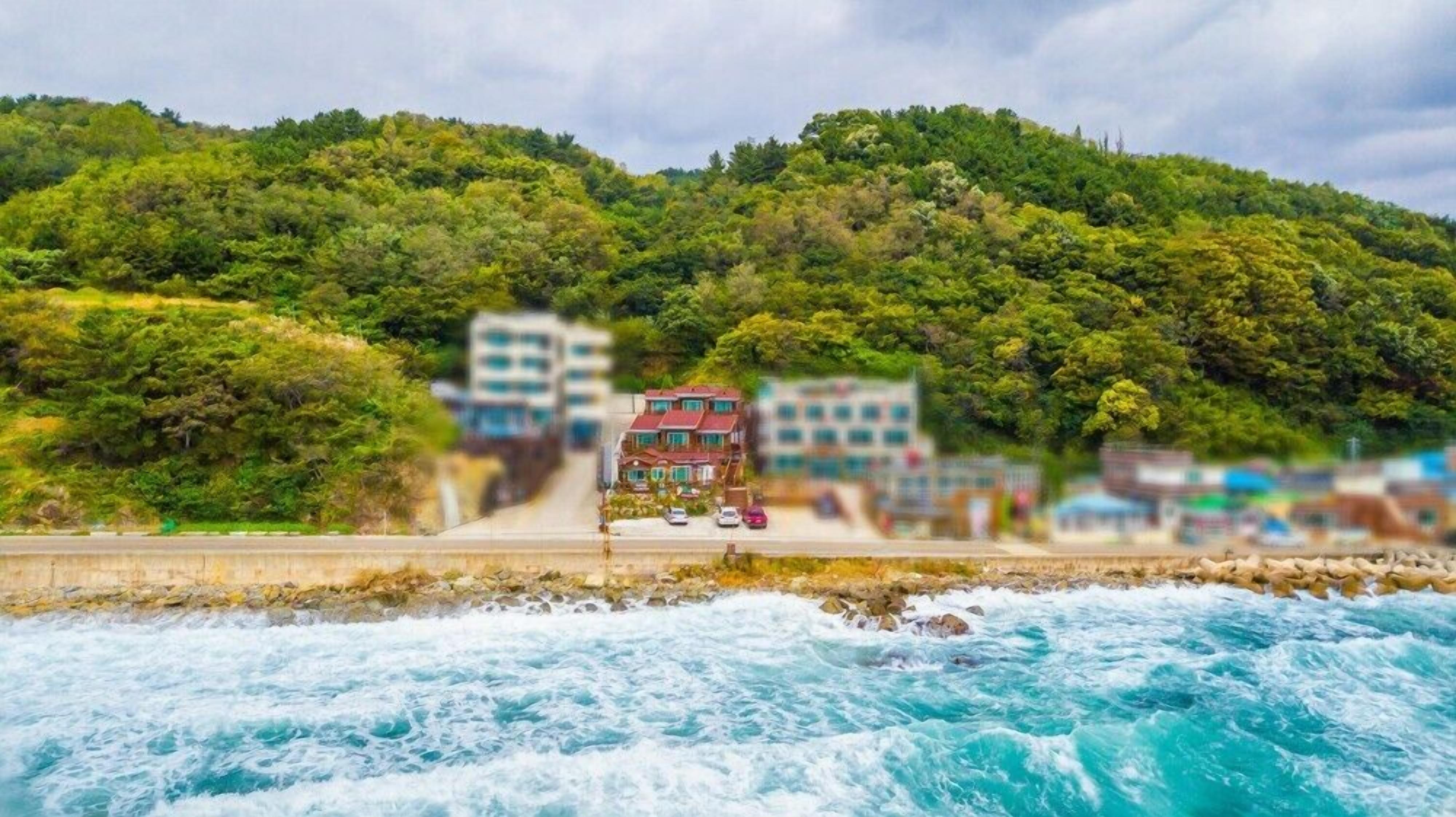 A coastal view with a building on a hillside, a road leading to it, and a rocky shoreline with waves.