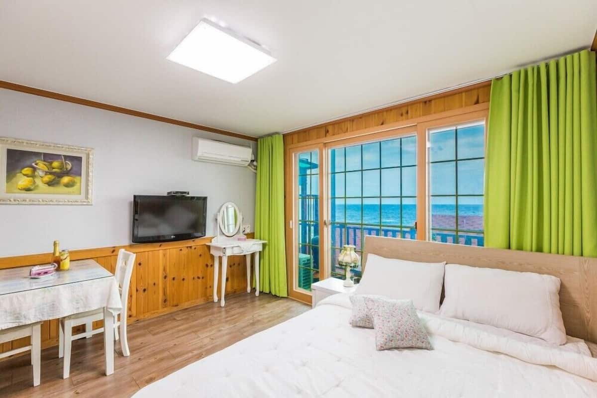 A bedroom with a bed, a desk, a television, and a view of the sea through large windows.