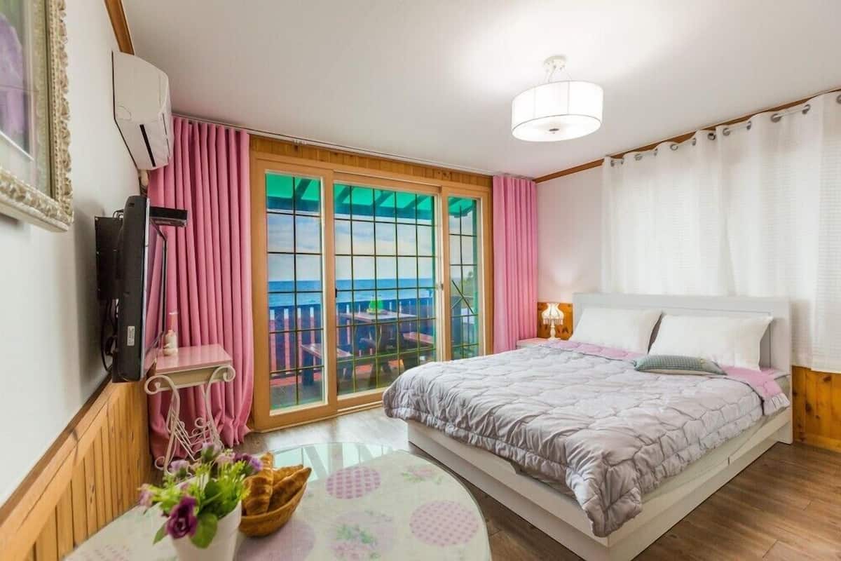 A bedroom with a bed, a TV, a window with curtains, a table with a vase, and a view of a balcony.