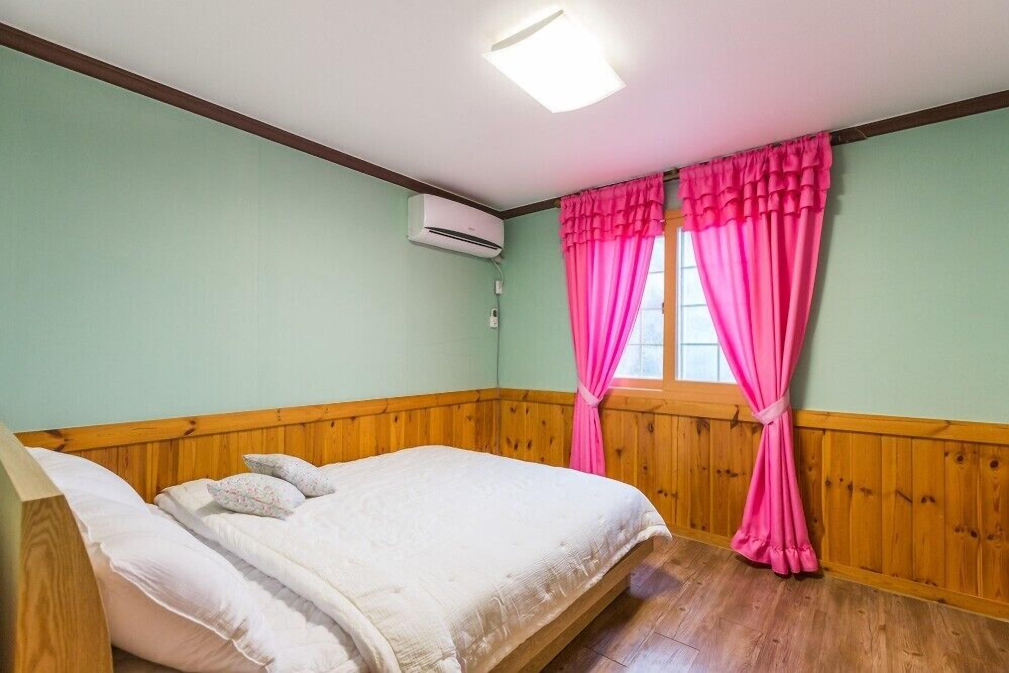 Un dormitorio con cabecera de madera, cama con ropa de cama blanca, ventana con cortinas rosadas y un equipo de aire acondicionado en la pared.