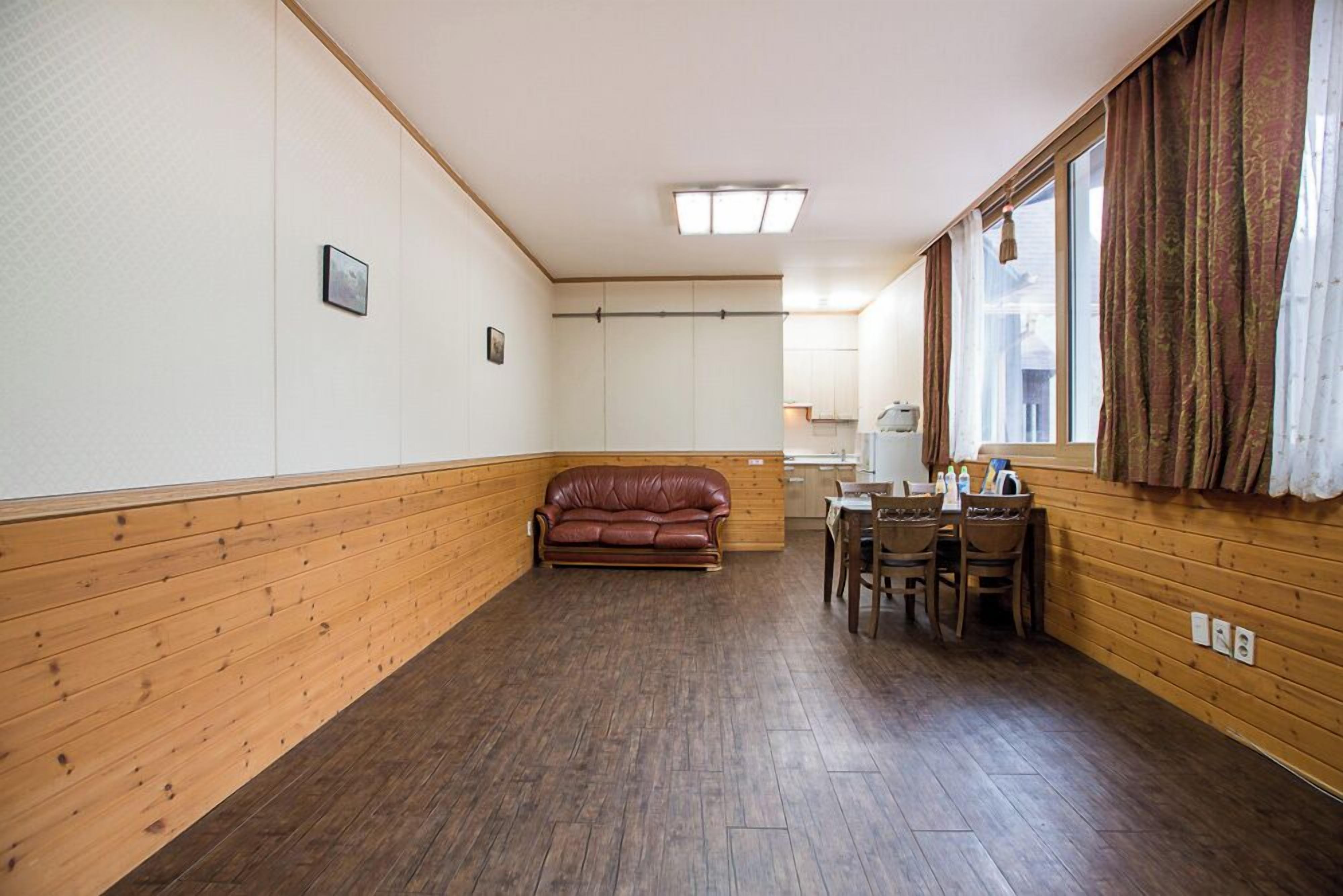 Una habitación con piso de madera, un sofá de cuero marrón, una mesa de comedor con sillas y una ventana con cortinas.