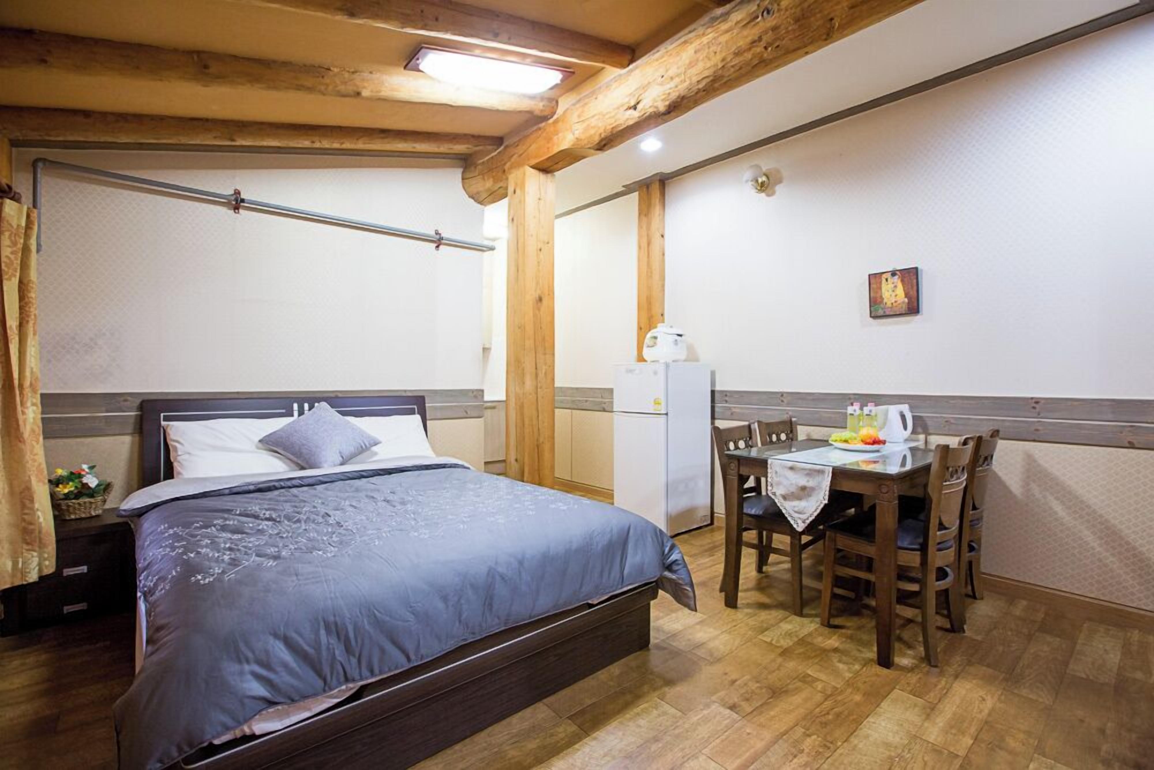 Un dormitorio con cama, una pequeña mesa con sillas, un refrigerador y una ventana con cortinas.