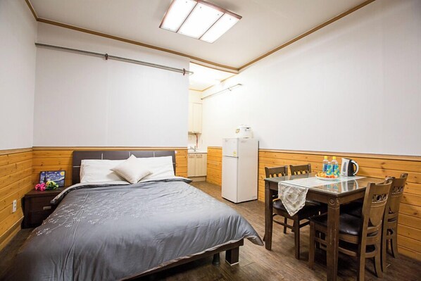 A bedroom with a bed, a small table with chairs, a refrigerator, and a wooden wall paneling.