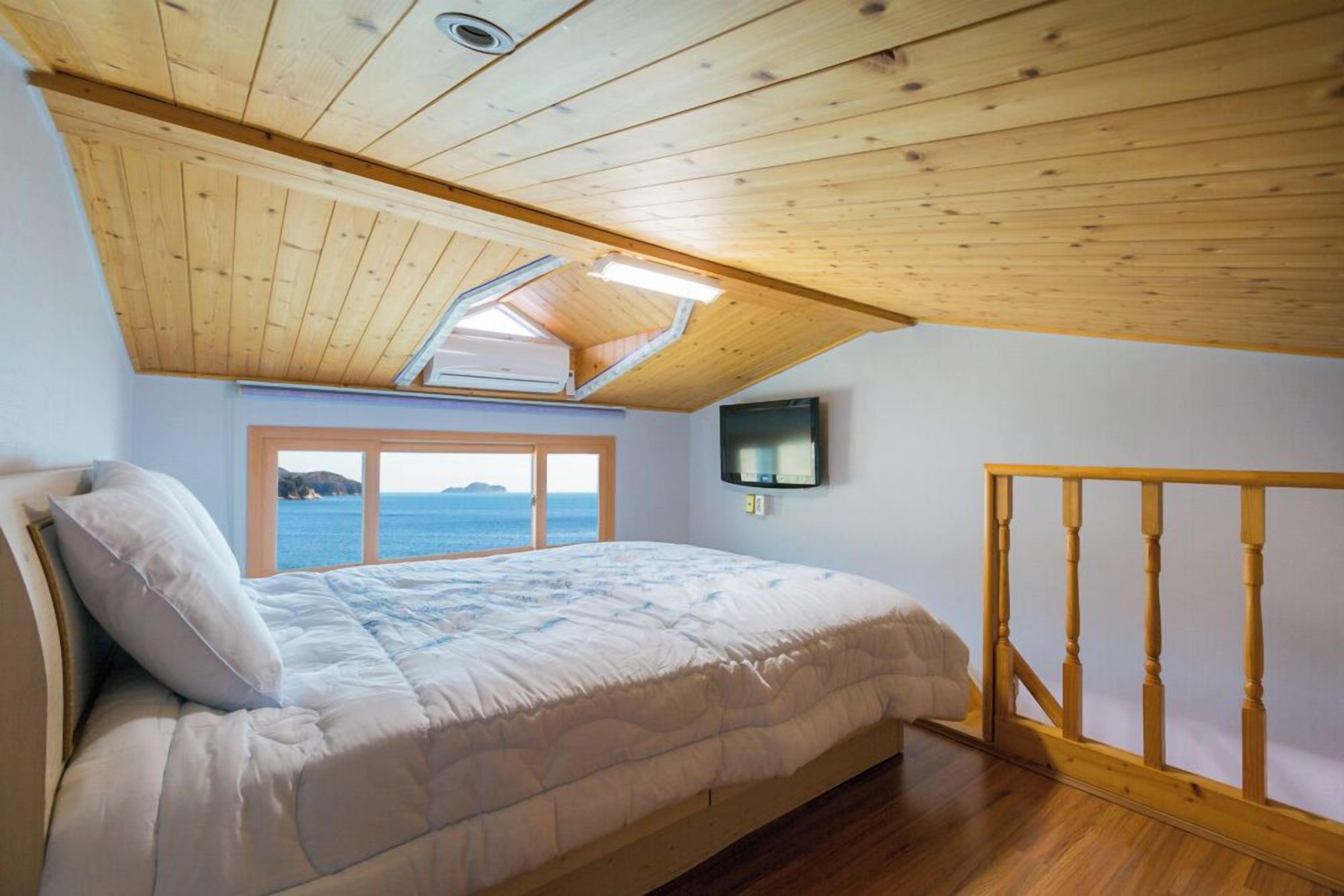 A bedroom with a wooden ceiling, a bed with white bedding, a window with a view of the sea, and a TV mounted on the wall.