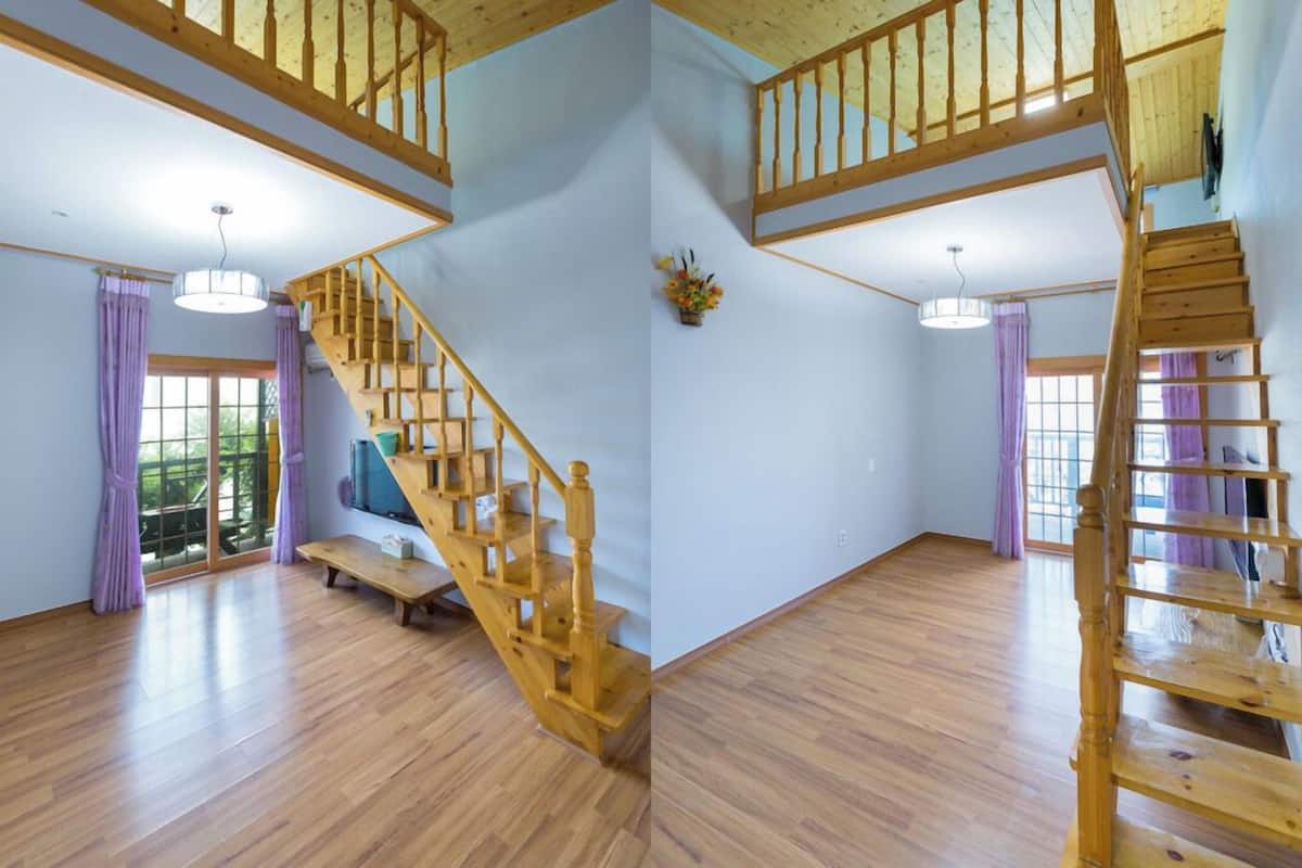Una habitación amplia con pisos de madera, una escalera con barandilla de madera y un ventanal con cortinas moradas.