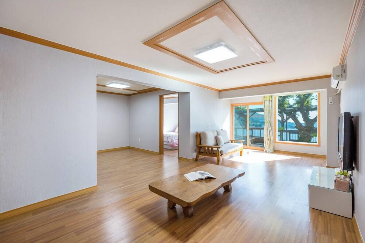 Una habitación amplia con suelo de madera, un ventanal con vistas, un banco de madera y un televisor en la pared.