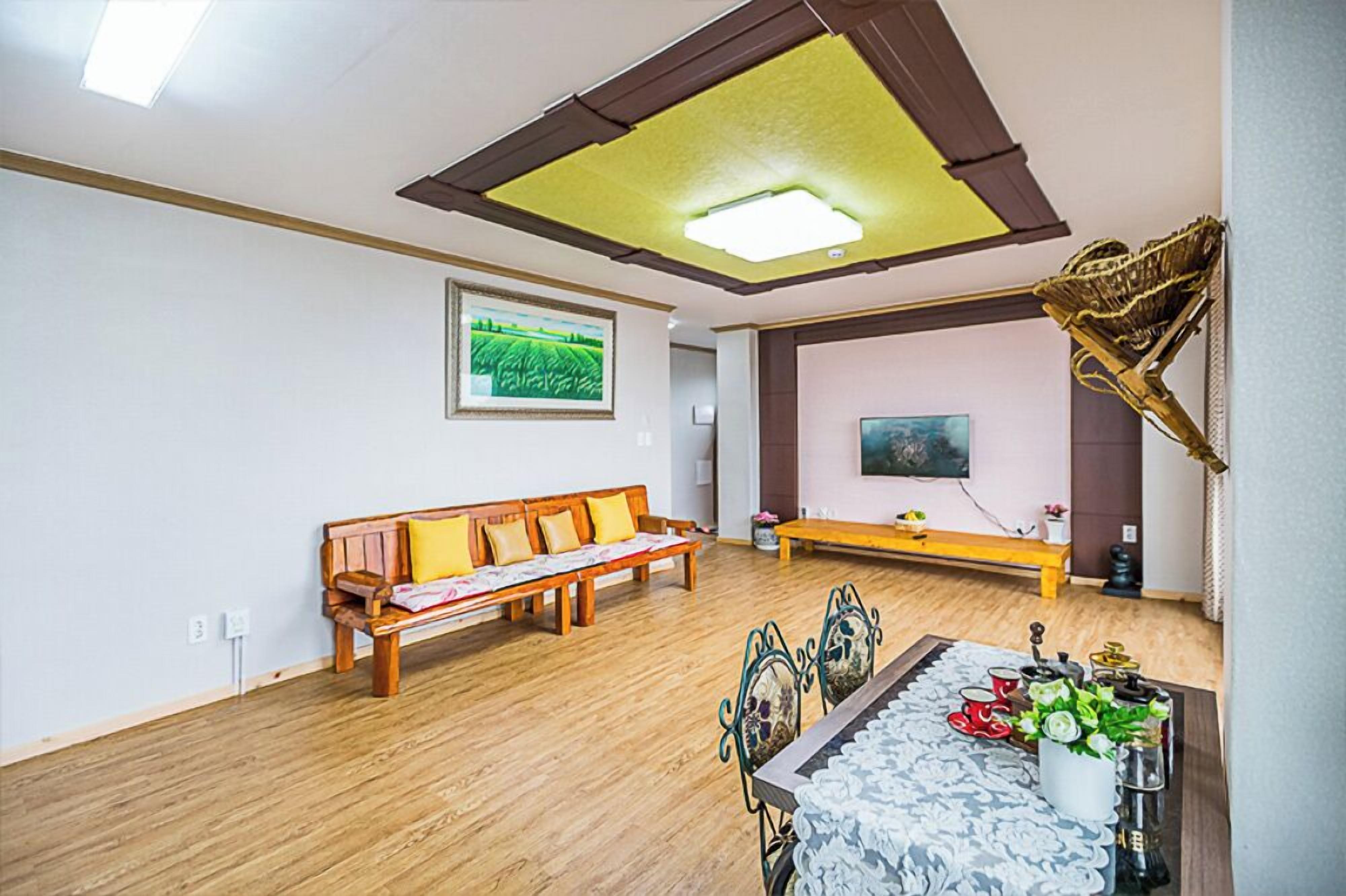 Quarto com um banco de madeira, uma mesa de jantar com cadeiras, uma luminária no teto e uma TV fixada na parede.