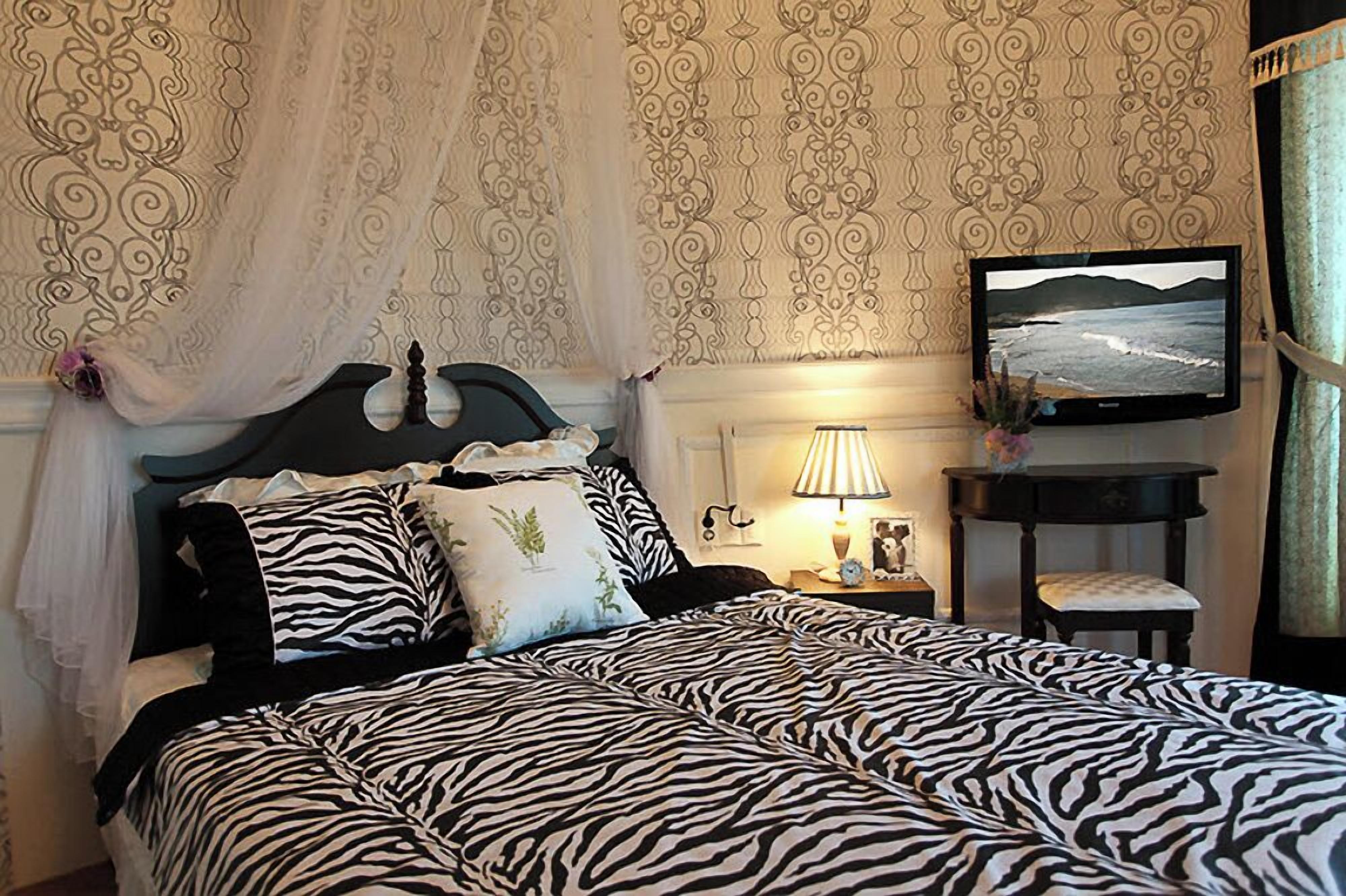 A bedroom with a zebra-patterned bedspread, a headboard with decorative elements, a bedside table with a lamp and a small plant, and a television mounted on the wall.