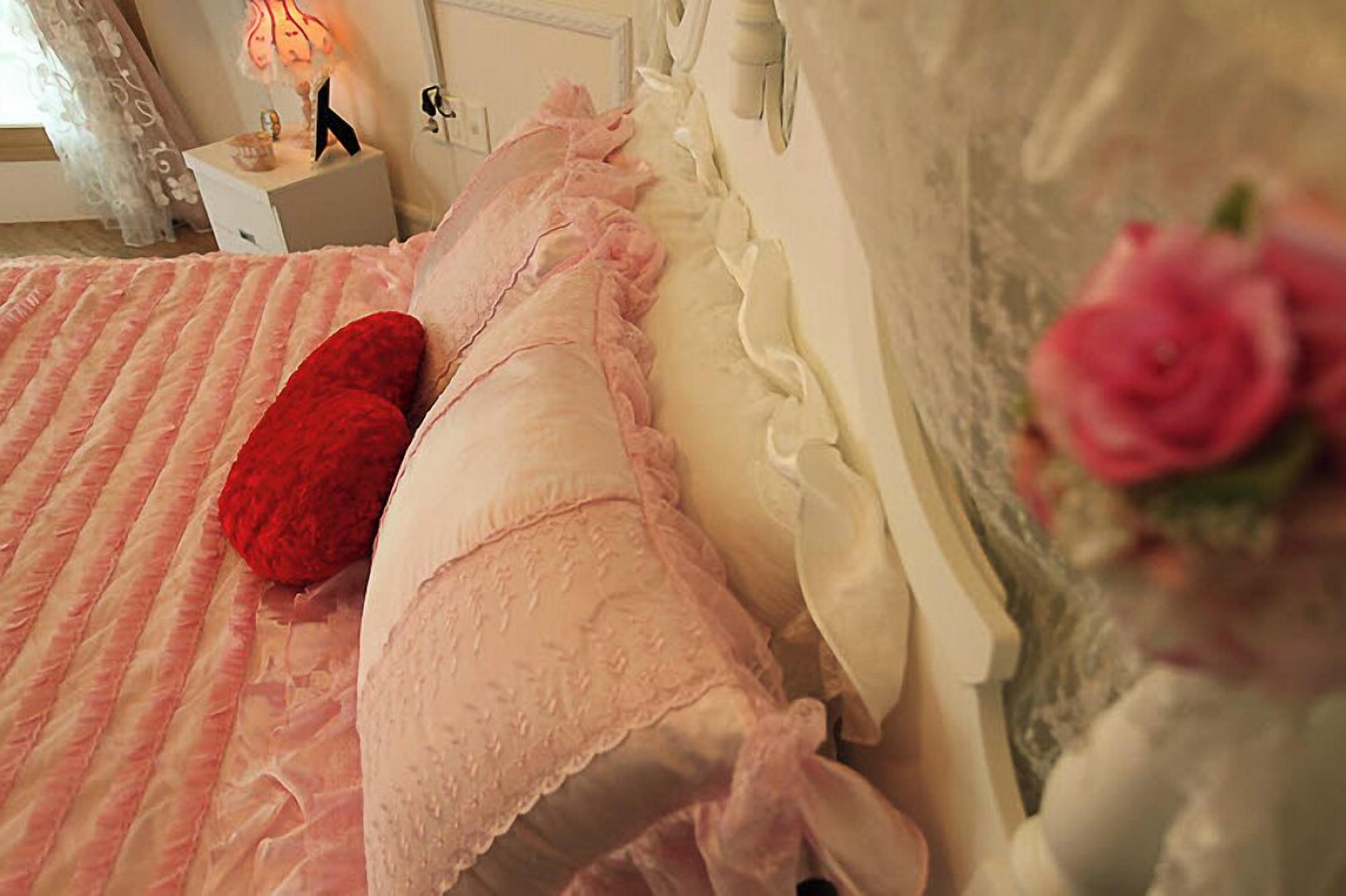 Una cama con ropa de cama rosa y una almohada roja, una mesita de noche con una lámpara, y una rosa rosa en un jarrón.