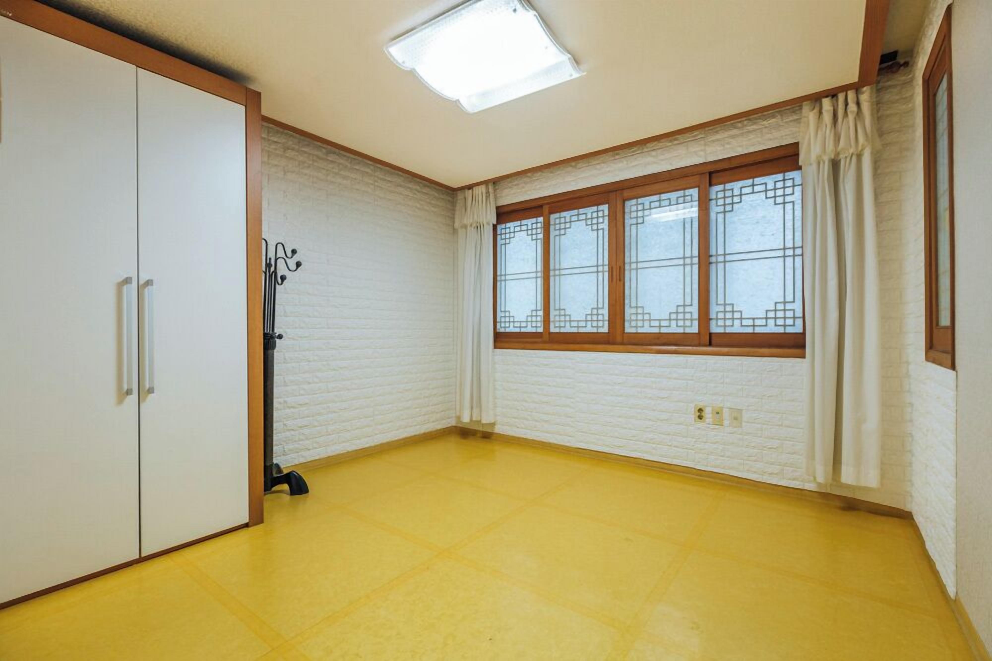 A room with a yellow floor, white brick walls, a large window with curtains, and a wardrobe with sliding doors.