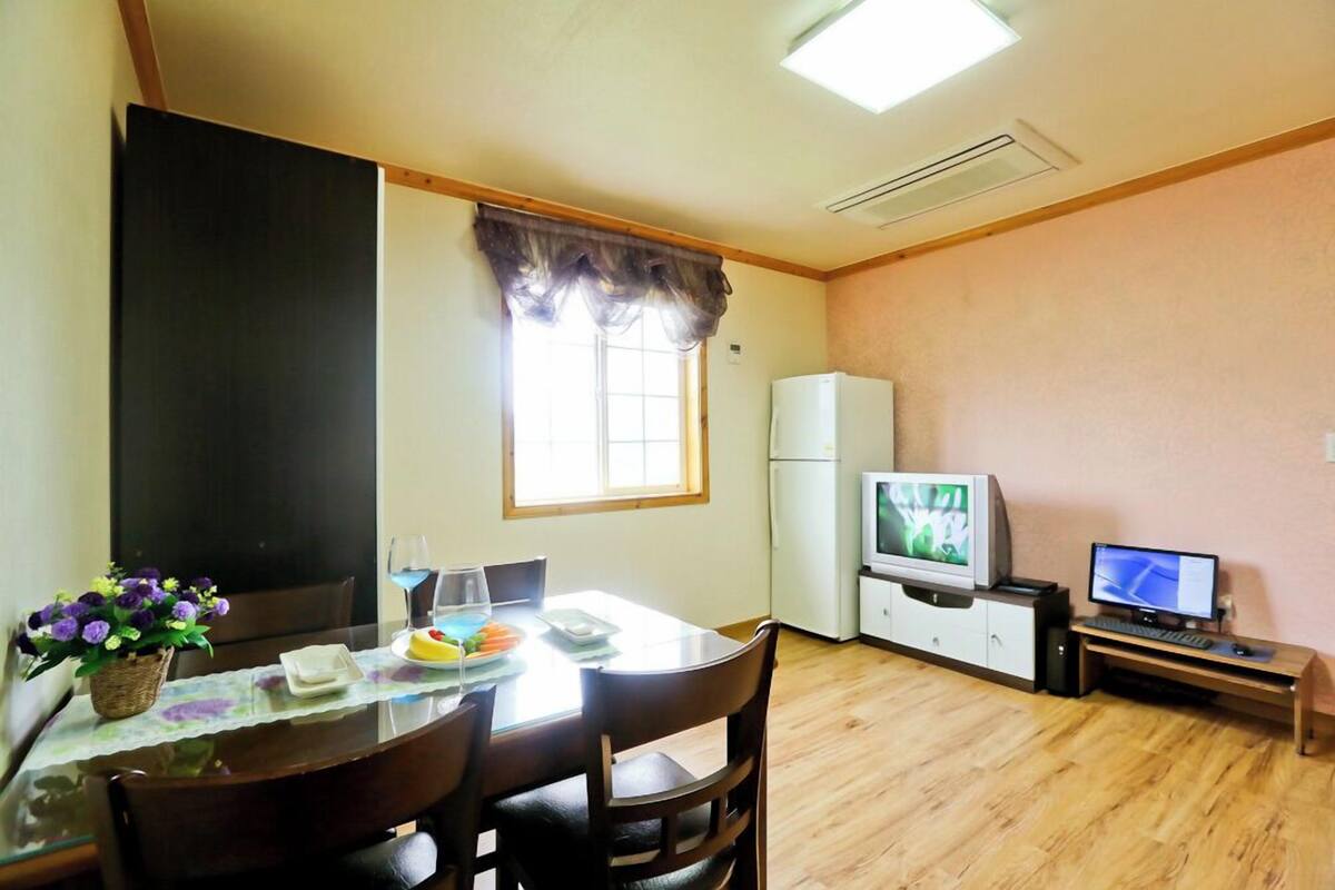 Un coin repas avec une table dressée pour deux personnes, une télévision, un ordinateur et un réfrigérateur en arrière-plan.