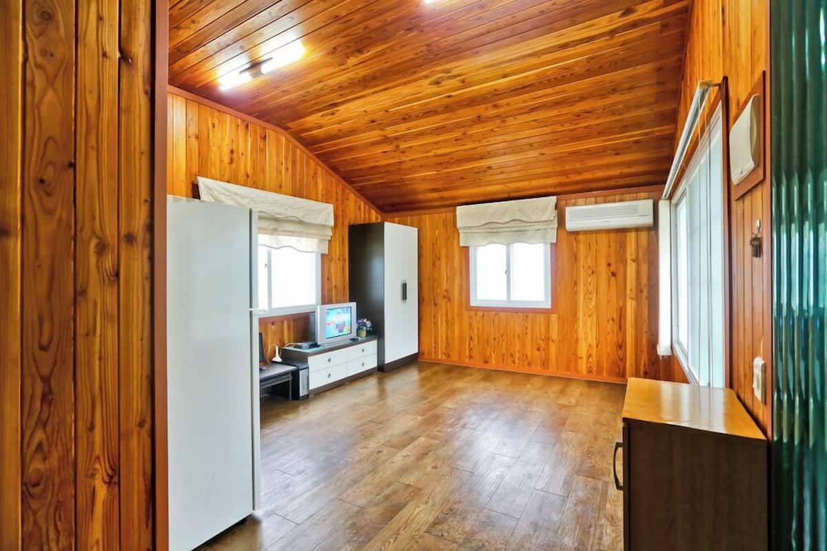 Une pièce spacieuse aux murs et au plafond en bois, avec un réfrigérateur blanc, une petite table sur laquelle se trouve un téléviseur, et une fenêtre avec des rideaux.