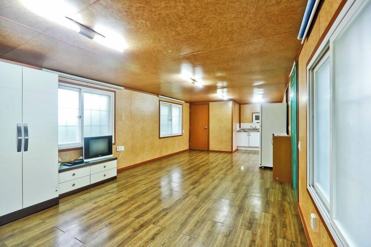 Une pièce spacieuse avec un plancher en bois, un téléviseur posé sur un meuble et un coin cuisine visible à travers une porte ouverte.