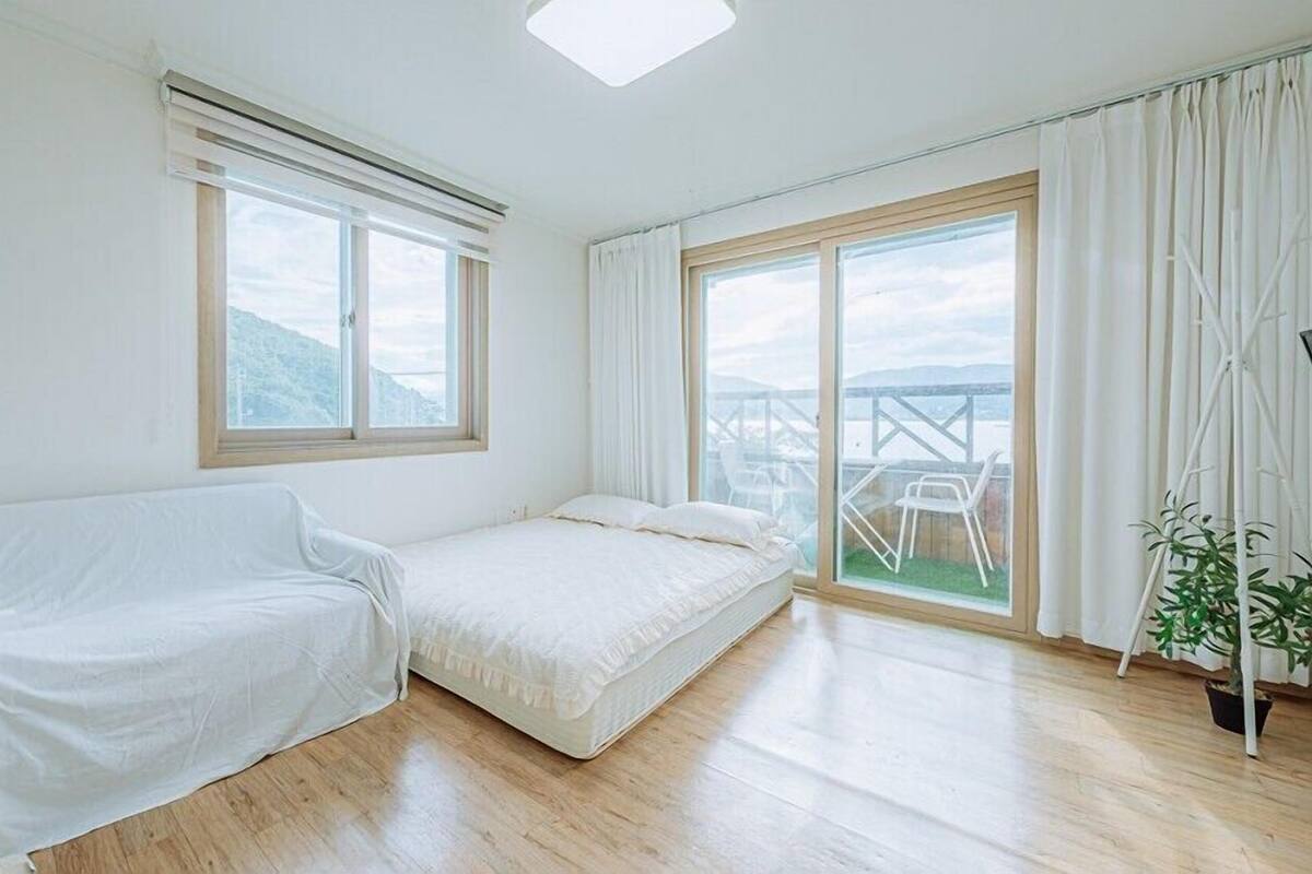 A room with a bed, a covered couch, a large window, and a view of a balcony with outdoor furniture.