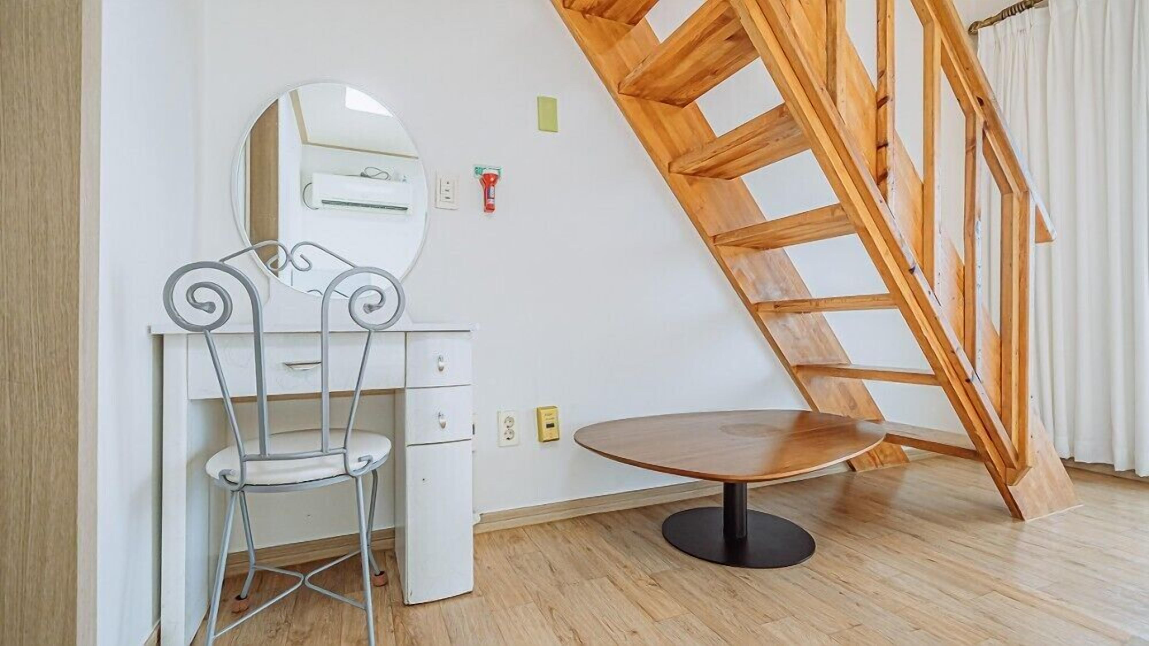 Una habitación con escritorio blanco, silla y mesa redonda, una escalera de madera y una pared blanca con espejo y enchufes.