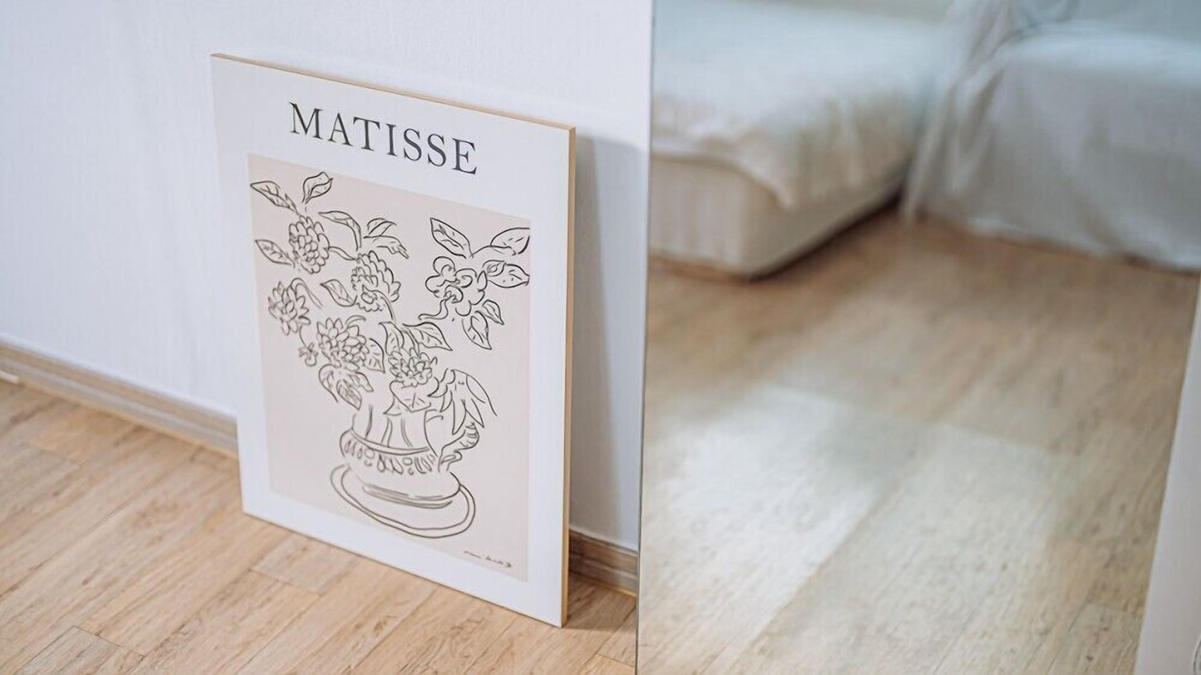 Una obra de arte enmarco titulada 'MATISSE' con un diseño floral, sobre un suelo de madera junto a una cama con ropa de cama blanca.