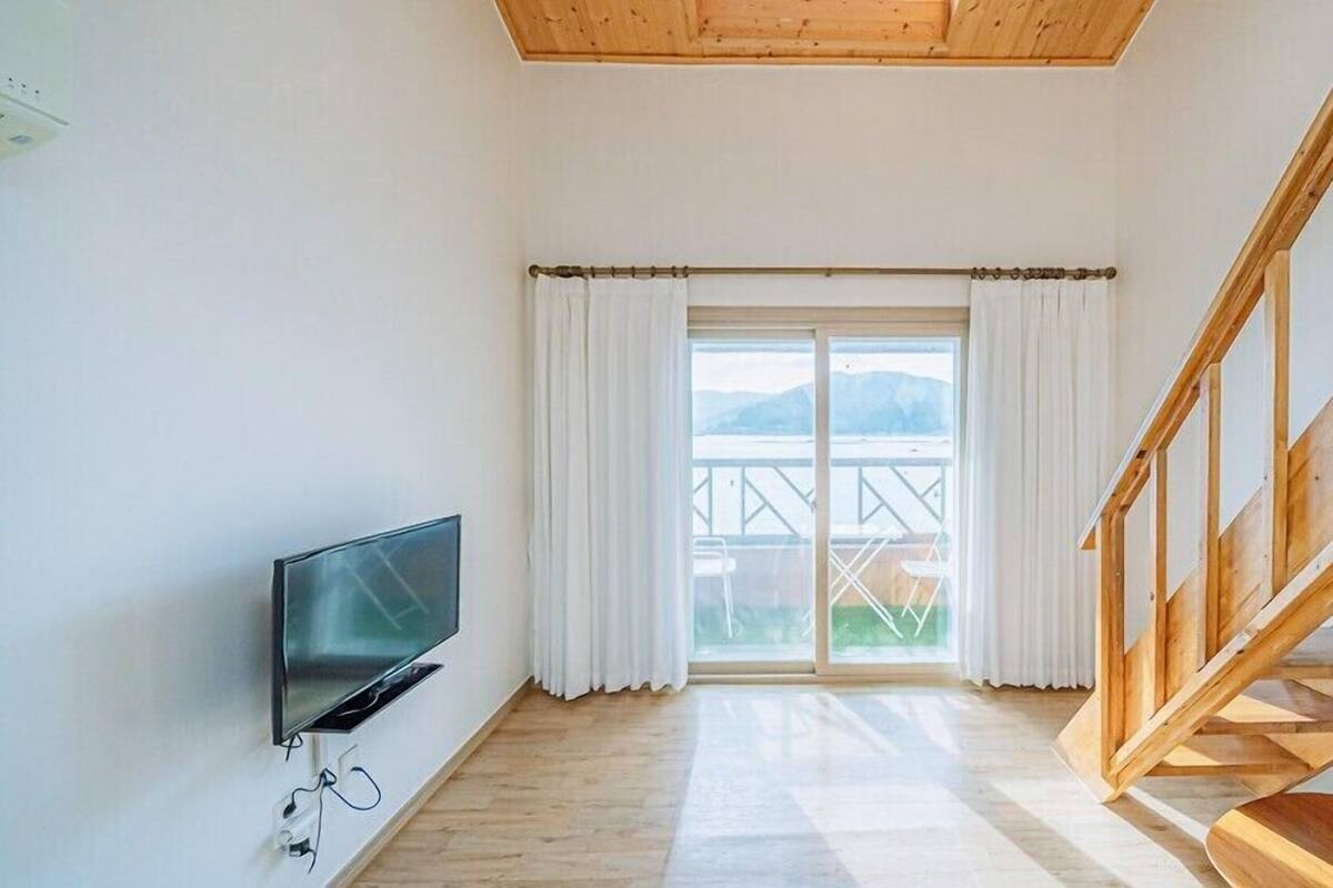 A room with a wooden staircase, a TV mounted on the wall, and a large window with curtains.
