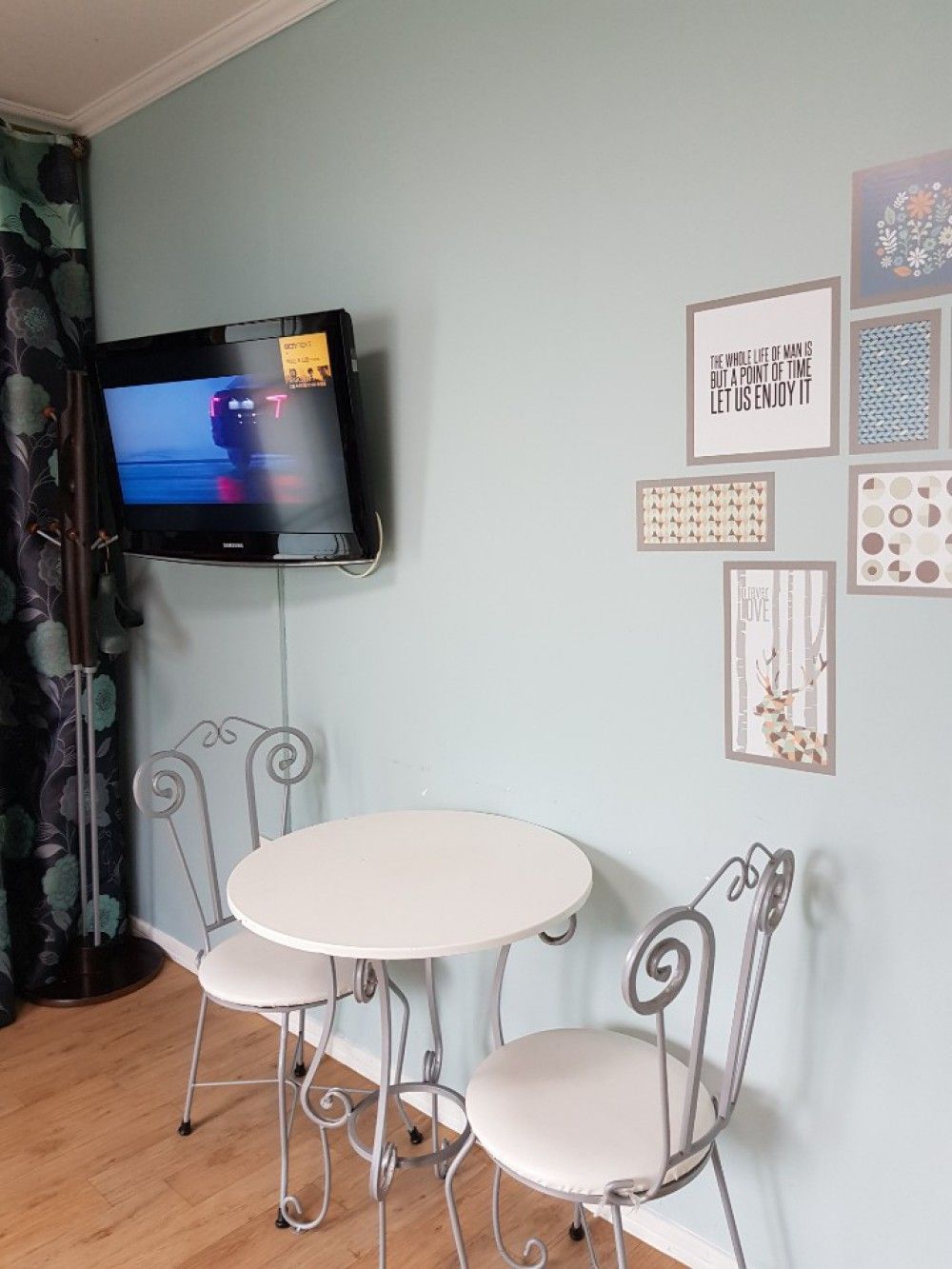 A room with a flat-screen TV mounted on the wall, a round table with two chairs, and various framed artworks on the wall.