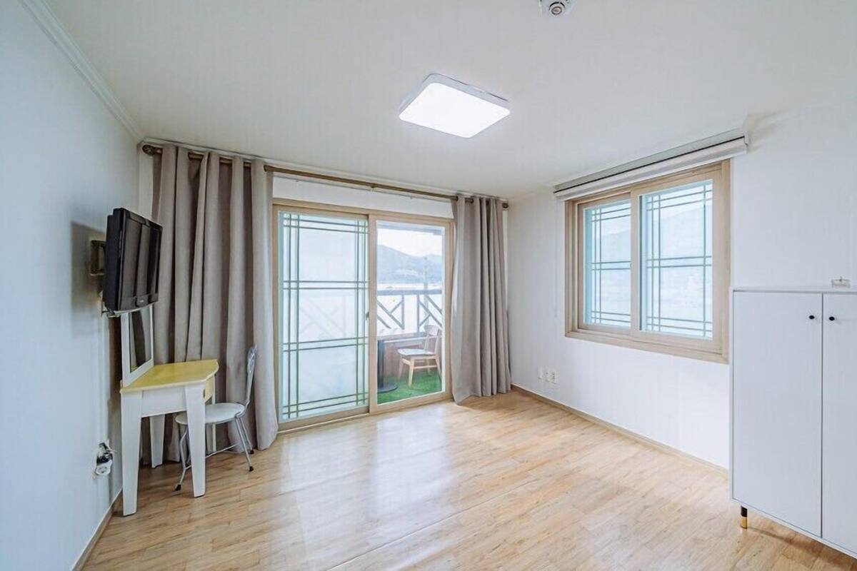 A room with a wooden floor, a white desk with a chair, a TV mounted on the wall, a sliding glass door leading to an outdoor area, and a white wardrobe.