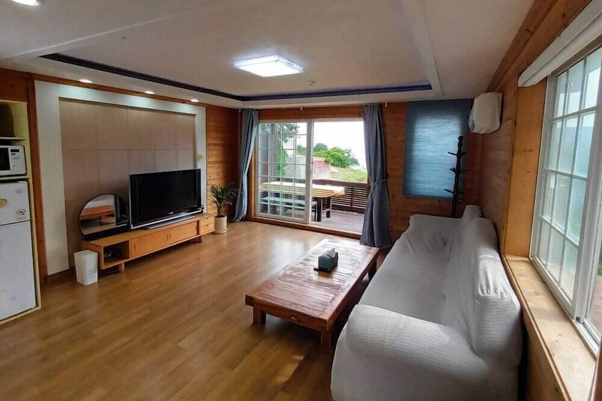 A modern living room with a wooden floor, a sofa, a coffee table, a TV, and a sliding glass door leading to an outdoor area.